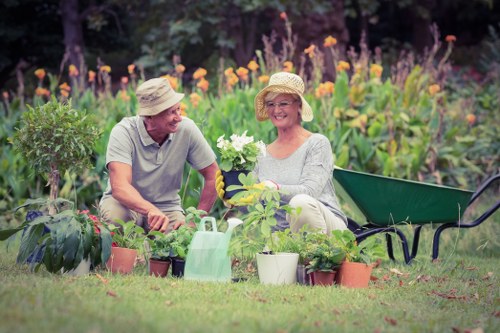 A middle-aged couple is gardening together in a lush, well-maintained backyard garden. The man, wearing a wide-brimmed straw hat and light grey T-shirt, is sitting on the grass, engaged in planting or tending to a green potted plant. The woman, also wearing a straw hat and casual light-colored clothing, is smiling and holding a white flowering plant, seated on a garden bench. Surrounding them are numerous plant pots with various young plants and garden tools, including a green watering can. The background features a vibrant flower bed filled with tall purple and orange blooming flowers, bordered by hedge or shrub borders, with lush green grass covering the lawn in the foreground. The scene is outdoors on a bright, sunny day, with natural light highlighting the vivid colours of the flowers and greenery, illustrating a peaceful and productive gardening activity typical of outdoor garden maintenance or planting projects in Kilburn or nearby areas.