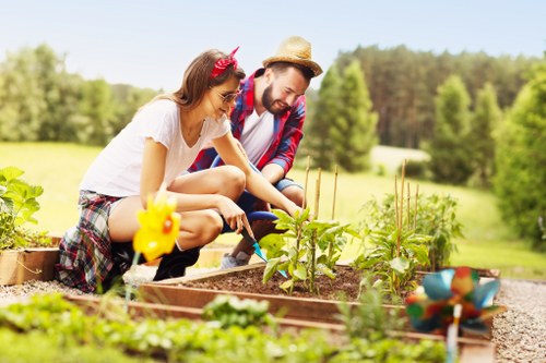 A young woman and a man are working together in a vegetable garden within a spacious outdoor area. The woman is crouching, wearing a white t-shirt and patterned shorts, with a pink headband, while the man, dressed in a plaid shirt and straw hat, is also kneeling beside her. They are tending to rows of young leafy plants, possibly vegetables, planted in well-structured raised garden beds made of wood. The garden features defined borders, with rich, dark soil surrounding the plants, and there are a few decorative garden ornaments visible, including small bird figures. In the background, a green lawn extends into a landscape of trees and shrubs, under a clear, bright sky, indicating pleasant weather. The scene’s natural tones of greens, browns, and vibrant plant colors reflect a healthy, cared-for outdoor space suitable for home gardening services offered by Landscapers Kilburn, serving the Kilburn area in London, NW6 postcode. The overall setting highlights active gardening in a well-maintained, peaceful garden environment, emphasizing the benefits of professional landscaping and garden maintenance services.