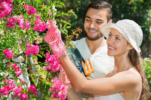In a well-maintained UK garden, a man and woman are engaged in gardening activities amidst lush greenery. The foreground features vibrant pink flowering bushes with dense clusters of blossoms and green foliage. The woman, wearing a white sun hat and gardening gloves, is tending to the plants with a small pruning tool, while the man, dressed in a white shirt, stands beside her, smiling and observing. The garden includes a neatly trimmed lawn and a variety of shrubbery and small trees in the background, with a mix of natural green tones and colorful blooms. The outdoor environment appears bright and sunny, highlighting the healthy, well-kept grass and the fresh, lively appearance of the garden's plant life. This scene exemplifies typical gardening activities and landscape features commonly found in residential outdoor spaces in Kilburn or similar London suburbs, reflecting the expertise of local landscapers such as Landscapers Kilburn who support garden maintenance and improvements.