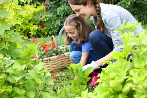 A woman and a young girl are gardening together in a lush outdoor space, surrounded by various green plants and shrubs. The girl is crouching down, holding a wicker basket filled with small red flowers, while the woman leans over beside her, smiling and assisting. The garden features a densely planted area with leafy bushes in the foreground and background, including a hedge and small flowering plants. The ground is covered with a well-maintained grass lawn, and parts of a paved patio are visible. The scene takes place in daylight with natural light illuminating the vibrant green foliage, suggesting mild weather conditions. This outdoor environment reflects typical features of a landscaped garden that might be maintained by local gardening services, such as those offered by Landscapers Kilburn in the Kilburn area, associated with the postcode NW6, highlighting the natural beauty and inviting atmosphere of a cared-for city garden.