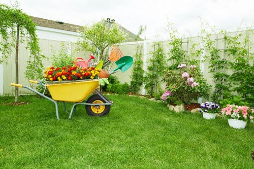 A well-maintained suburban garden in Kilburn featuring a vibrant green lawn with evenly cut grass, bordered by a white privacy fence covered in climbing plants and vines. In the foreground, a yellow wheelbarrow filled with colourful garden flowers and gardening tools sits on the grass, suggesting ongoing garden maintenance. To the right, there are potted flower beds with a variety of blooming plants, including pink and purple blossoms, arranged in front of a cluster of shrubs and small trees. The background reveals a few garden trees and a partly cloudy sky, indicating mild weather conditions. The scene reflects a neat and inviting outdoor space suitable for gardening services focused on lawn care, plant care, and general garden tidying in Kilburn, which landscapes and maintains outdoor areas for quality garden enjoyment.