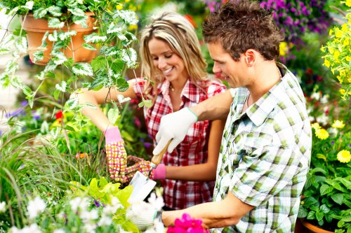 A gardener and a women working together in a lush garden with vibrant flowers, verdant shrubs, and well-maintained lawn. The scene shows a paved pathway bordered by flower beds containing a variety of colourful blooms and green foliage, with a wooden deck partially visible in the background. The garden features a mix of dense shrubbery, potted plants, and small trees, contributing to a tidy and cultivated outdoor space typical of residential landscaping in Kilburn. Bright natural light suggests a clear, sunny day, highlighting the rich textures and colours of the garden elements. The overall environment presents a healthy, flourishing outdoor area suitable for garden maintenance, planting, and landscape enhancement tasks, reflective of professional gardening services offered by Landscapers Kilburn.