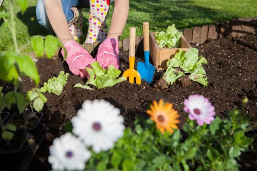 In the foreground of the garden, vibrant flower beds feature colourful blooms including white, orange, and purple flowers, adding visual interest to the outdoor space. Behind the flowers, a neatly maintained lawn with dense, green grass extends towards a flower bed border lined with small plants. The middle ground shows a child wearing pink gardening gloves, planting young lettuce or similar leafy greens into rich, dark soil using small wooden garden tools such as a trowel and rake, which are positioned upright in the soil. The garden area is part of a well-organised outdoor space, likely part of a residential garden in Kilburn, with a clear separation between the flower beds, lawn, and vegetable patch. The scene appears to be under bright natural sunlight, with a lush, green backdrop of trees or shrubbery suggesting a peaceful, well-tended garden environment suitable for gardening and landscaping services from Landscapers Kilburn.