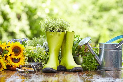 The image displays a garden scene with a weathered wooden table holding garden tools and decorative items. Two bright green rubber gardening boots are positioned upright with their openings filled with small white flowering plants, placed centrally on the table. To the left, there is a cluster of vibrant yellow sunflowers and other mixed flowers, adding natural colour contrast. On the right, a metal watering can with a long spout sits beside a green plastic plant pot, which contains additional green plants. The scene is set outdoors, with a blurred background of lush, green foliage and trees, indicating a well-maintained garden environment. The surface of the table appears slightly wet, suggesting recent watering or rain, and the overall lighting is natural, compatible with a mild, cloudy day. This setup subtly reflects outdoor gardening activities typical for a residential garden in Kilburn or surrounding areas, aligning with professional gardening services offered by Landscapers Kilburn.