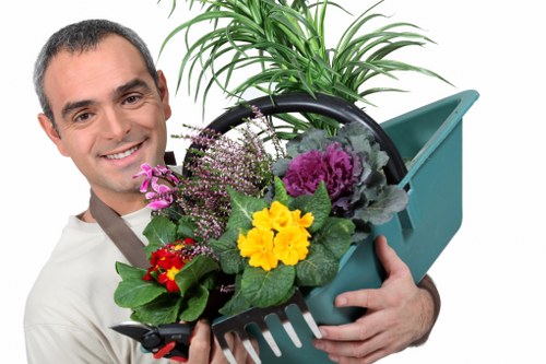 The image depicts a smiling man holding a turquoise gardening tool bucket filled with a variety of colorful plants and flowers, including yellow, purple, and red blossoms, alongside lush green foliage. Behind him is a tall, healthy green plant with broad leaves, suggesting a well-maintained garden environment typical of outdoor spaces in Kilburn. The background consists of a plain white area, focusing attention on the gardening tools and plant arrangements, indicative of professional gardening or landscaping services offered by Landscapers Kilburn. The scene conveys the freshness, vitality, and careful plant selection associated with garden renovation ideas, highlighting the importance of garden planting, planting supplies, and outdoor plant care in creating vibrant, inviting outdoor spaces. Such detailed and natural plant displays and tools align with services in lawn and garden care, reflecting the quality and variety available for garden improvement projects in the Kilburn area.
