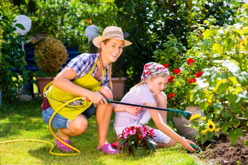 In a lush, well-maintained garden, a woman and a young girl are engaged in outdoor gardening activities. The woman is crouched down, smiling, and wearing a straw hat, a yellow apron, and casual clothing suitable for gardening. She appears to be helping or supervising as the girl, dressed in a light-colored outfit with a floral hat, tends to a flower bed with colorful blooming flowers, including reds, yellows, and pinks. The garden features a neat grassy lawn, bordered by flower beds and hedges, with a variety of plants and shrubs creating a vibrant green background. Paving stones or a small patio area can be seen nearby, providing a clean surface for outdoor activities. There are garden lights and lush foliage, suggesting an inviting and cared-for outdoor space, likely part of a residential garden in Kilburn or the surrounding London area. The scene captures a moment of outdoor family gardening, emphasizing natural textures, bright natural daylight, and the natural tones of plants and flowers. This setting highlights the landscape gardening services offered by Landscapers Kilburn to create vibrant, functional outdoor spaces for families in the local area.