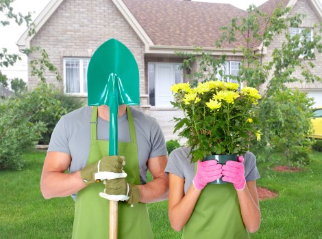 A garden scene featuring two individuals standing on a well-maintained lawn in front of a suburban house with grey brick walls and white window frames. One person, wearing a light grey t-shirt, green gardening gloves, and a green apron, holds a blue gardening spade with the blade resting on the ground. The other person, dressed in a grey t-shirt and pink gardening gloves, holds a large potted plant with vibrant yellow flowers and lush green foliage close to their face. The garden includes neatly trimmed bushes and small trees, with a backdrop of a paved pathway and a yellow vehicle partially visible on the right. The house behind them has a pitched roof, and the overall scene suggests outdoor lawn and garden maintenance, typical of gardening services in Kilburn. The outdoor environment appears bright and slightly overcast, indicating a mild weather day suitable for gardening work. This image underscores landscaping and lawn care activities that are offered by Landscapers Kilburn in the local area.