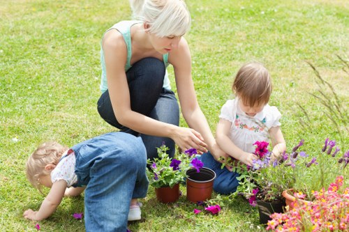 A woman and two young children are engaged in gardening activities in a well-maintained backyard garden, which features a lush, evenly cut grass lawn and a variety of flowering plants in pots and beds. The woman, with short blond hair, is kneeling on the grass, focused on planting or tending to purple and pink flowering plants in pots, while the children, dressed in casual clothing, assist by exploring the flower beds and soil around them. The garden background includes a clean, green lawn with a boundary hedge or shrubbery, suggesting an enclosed outdoor space suitable for family gardening. The scene appears to be during daylight with bright, natural lighting, highlighting the vibrant colours of the flowers and the natural tones of the garden environment. This setting reflects a typical outdoor space maintained by professional landscapers such as Landscapers Kilburn, integrating garden planting with family activities in a safe, inviting manner suitable for local garden care and outdoor maintenance services.