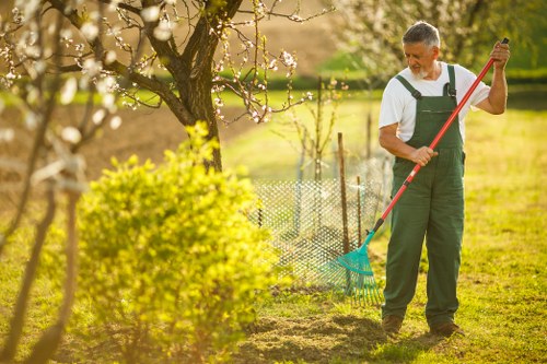 A man wearing green overalls and a white shirt is working in a backyard garden, using a rake to tend to a neatly cultivated patch of soil. The garden features a lush, well-maintained lawn with vibrant green grass and a variety of plants, including a flowering tree with pink blossoms in the background and a neatly trimmed hedge border. To the right, there is a small, colorful fence enclosing a section of garden beds, and a wooden deck is partially visible in the distance. The scene is captured on a bright, sunny day with natural light illuminating the outdoor space, indicating seasonal springtime conditions. The setting reflects professional garden maintenance services offered around Kilburn and the surrounding areas, emphasizing landscaping and planting work typical of local gardening projects managed by Landscapers Kilburn.